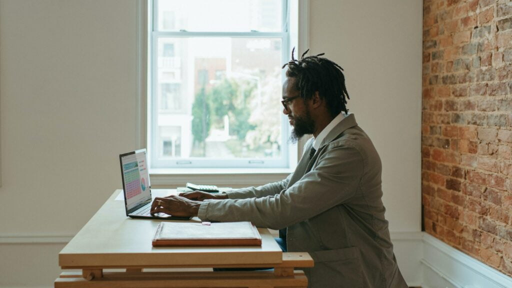 A man sitting at a wooden desk, working on a laptop in a room with a large window and a brick wall. He is wearing glasses and dressed in a suit, suggesting a professional setting. His attention is focused on the screen of his laptop, which is facing away from the camera, making it impossible to see what's on the screen. The room appears bright and airy, with natural light coming in from the window. The man's posture is relaxed yet attentive, indicating he is engaged in his work. There are a few items on the desk, including what appears to be a large book or folder and a calculator, contributing to the office-like environment.