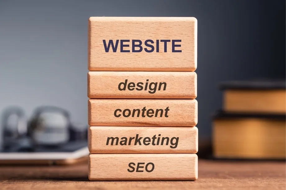 a set of wooden blocks stacked on top of each other, similar to a tower. Each block is inscribed with a different word associated with website creation and management. From top to bottom, the words are: WEBSITE design content marketing SEO The stack appears to represent the building blocks of a successful website, with SEO (Search Engine Optimization) as the foundation, followed by marketing, content, and design, and topped with the website itself. The blocks are situated on a table, and there is a blurred background featuring what appears to be a room with books to the right side, suggesting an office or study environment. The focus is on the blocks, which are in sharp contrast to the softly blurred background.